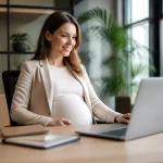 Nėščia moteris dirba prie kompiuterio moderniame biure, šypsosi